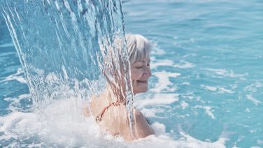 Senior caucasian smiling woman with gray hair enjoying falling on her shoulders flow of water. Hydromassage in outdoor thermal swimming pool. Active elderly concept. Blurry movement